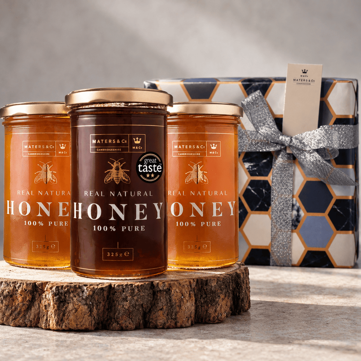 Three glass jars of MATERS & CO. honey displayed on a rustic wood slice, with a branded gift box in the background.