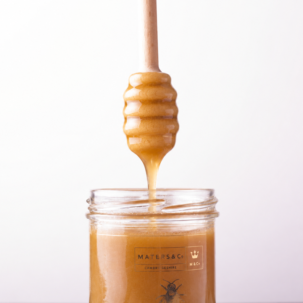 A wooden honey dipper dripping thick scottish heather honey into a clear glass jar labeled Maters & Co, set against a clean white background.